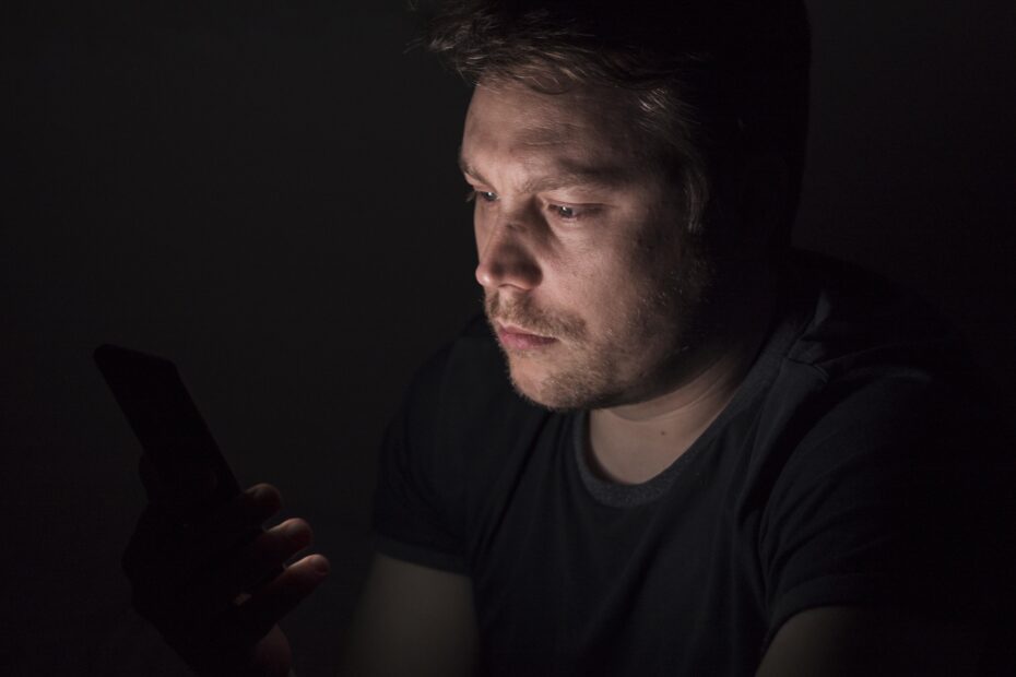 O rosto de um homem branco aparece por meio da luminosidade gerada apenas por um celular. O fundo da imagem é preto.