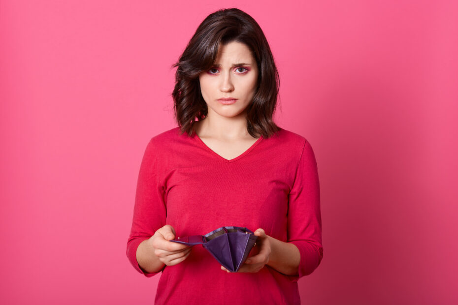 Mulher branca, dos cabelos pretos, usa uma camiseta cor de rosa e tem uma carteira aberta e vazia nas mãos. Ao fundo, uma parede cor de rosa.