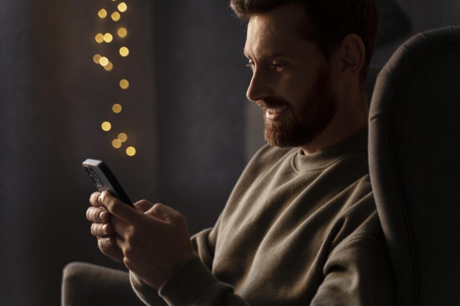 Homem branco, sentado em um sofá com o celular na mão, de onde saem ícones lumisos, aparece de lado enquanto olha atentamente para o celular.