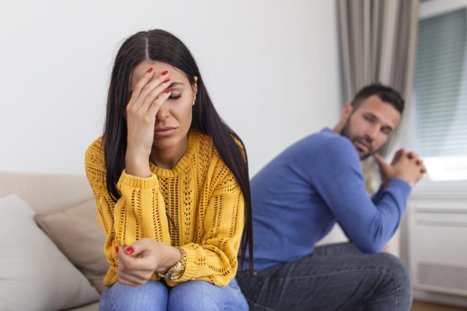 Mulher jovem com expressão de tristeza e frustração sentada em um sofá, usando suéter amarelo e cobrindo parte do rosto com a mão. Ao fundo, um homem com semblante sério e braços apoiados nas pernas a observa.
