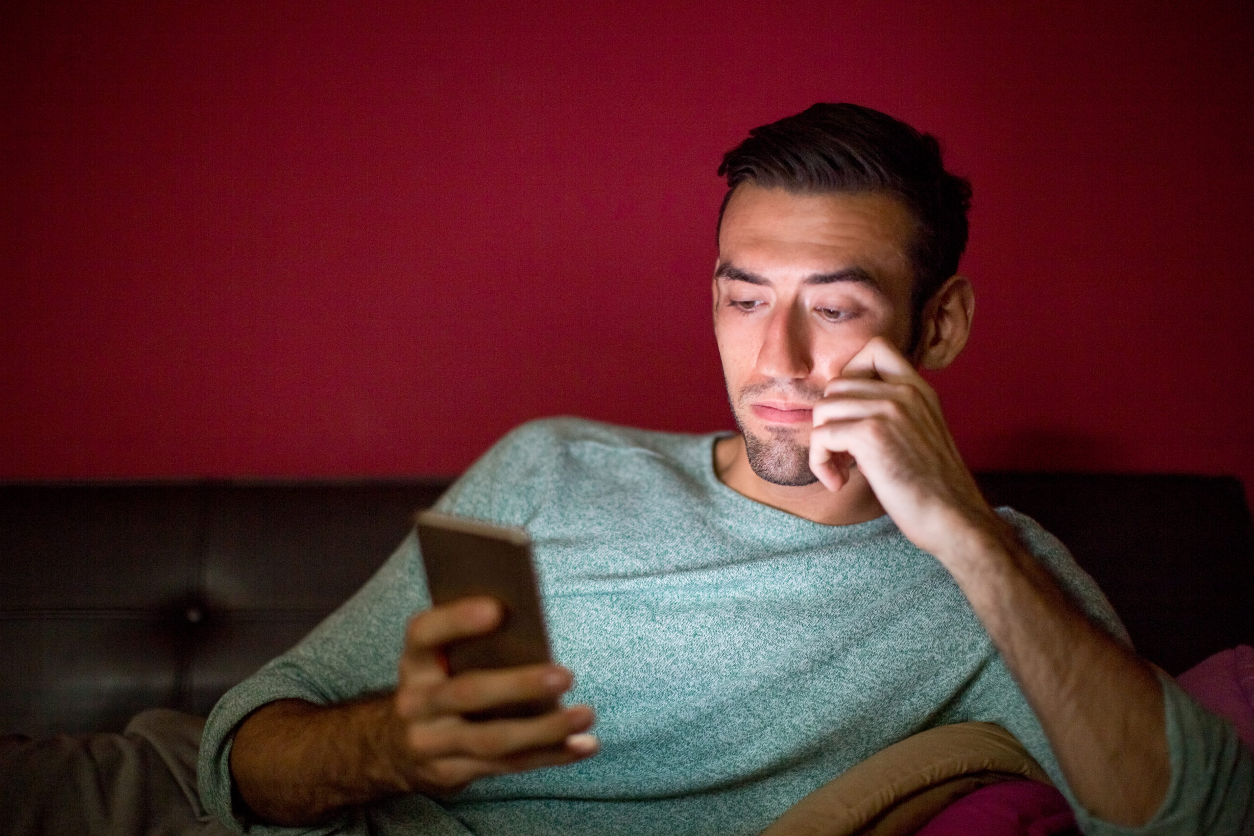 Homem sentado ao sofa olhando para o celular. Ao fundo, uma parede vermelha.