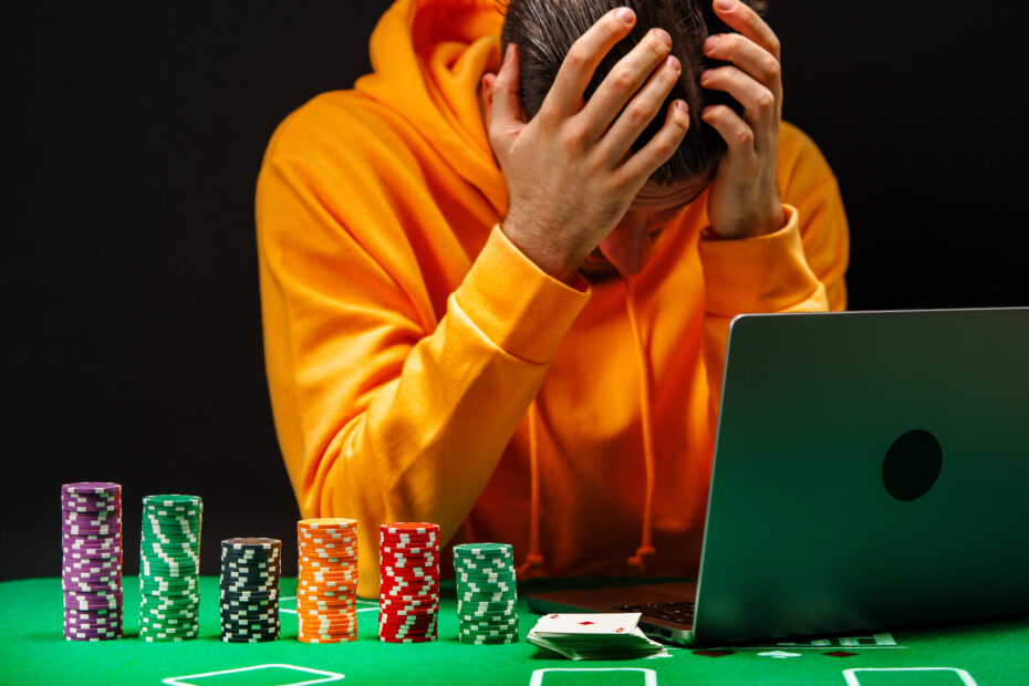 Homem com expressão de frustração e angústia, vestindo moletom laranja, segura a cabeça com as mãos enquanto olha para um notebook. À sua frente, sobre uma mesa verde, há várias pilhas de fichas de pôquer e cartas, sugerindo um cenário de jogos de azar online.
