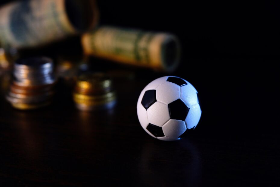 Mini bola de futebol sobre uma superfície escura, com moedas empilhadas e cédulas enroladas desfocadas ao fundo. A imagem simboliza a relação entre dinheiro e futebol, sugerindo temas como apostas esportivas ou investimentos no esporte.
