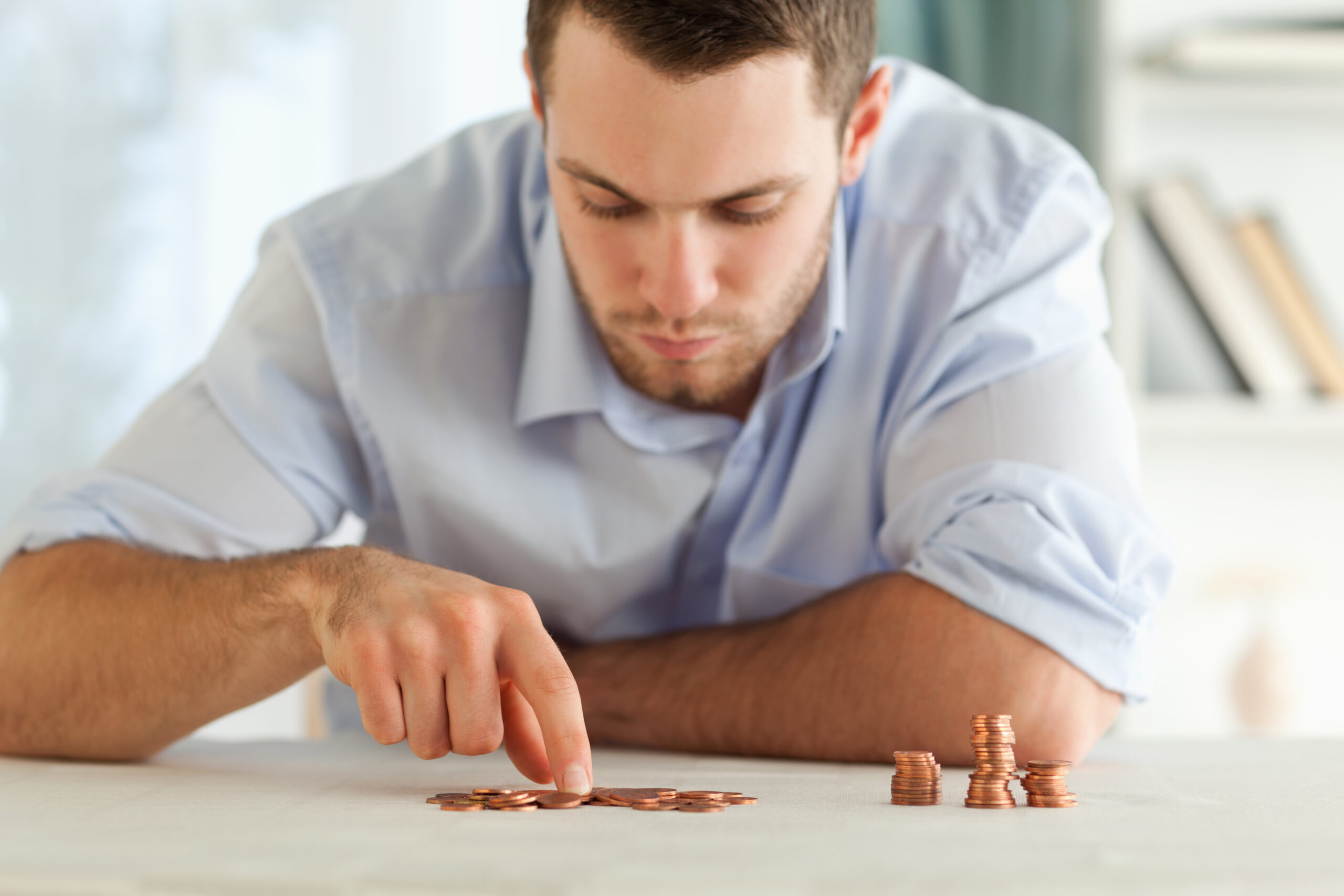 Homem de camisa azul clara observa com atenção várias moedas empilhadas sobre uma mesa, enquanto separa outras com o dedo. A cena transmite concentração e preocupação com finanças ou controle de gastos.