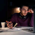 Homem jovem, usando fones de ouvido no pescoço e suéter roxo, trabalha concentrado à noite diante de um laptop. Ele segura uma caneta e um caderno, com documentos espalhados sobre a mesa e uma xícara de café ao lado.