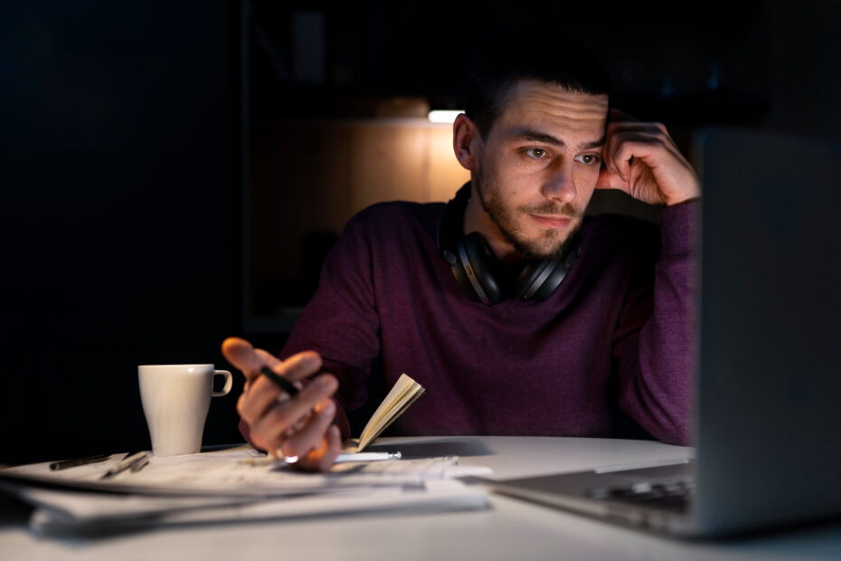 Homem jovem, usando fones de ouvido no pescoço e suéter roxo, trabalha concentrado à noite diante de um laptop. Ele segura uma caneta e um caderno, com documentos espalhados sobre a mesa e uma xícara de café ao lado.