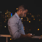 Homem de camisa social clara, apoiado em uma varanda à noite, usando o celular. Ao fundo, a cidade iluminada com luzes de prédios e postes.
