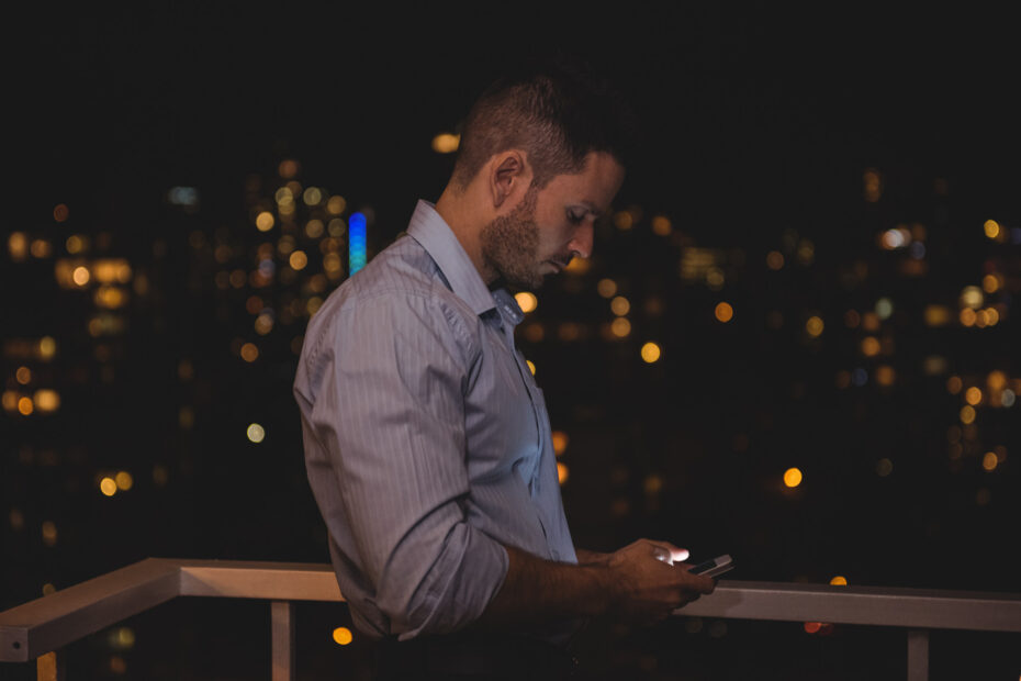 Homem de camisa social clara, apoiado em uma varanda à noite, usando o celular. Ao fundo, a cidade iluminada com luzes de prédios e postes.