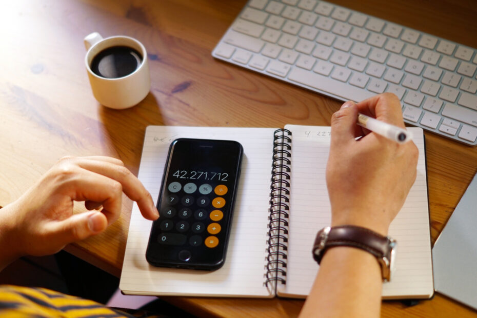 Pessoa usando um celular com aplicativo de calculadora aberto, anotando em um caderno ao lado de um teclado de computador e uma xícara de café, sobre uma mesa de madeira.