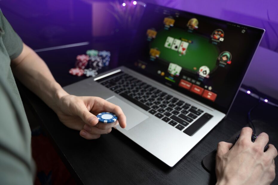 Pessoa segurando uma ficha de pôquer em frente a um notebook com jogo de pôquer online na tela, em um ambiente escuro com iluminação violeta e pilhas de fichas ao fundo.