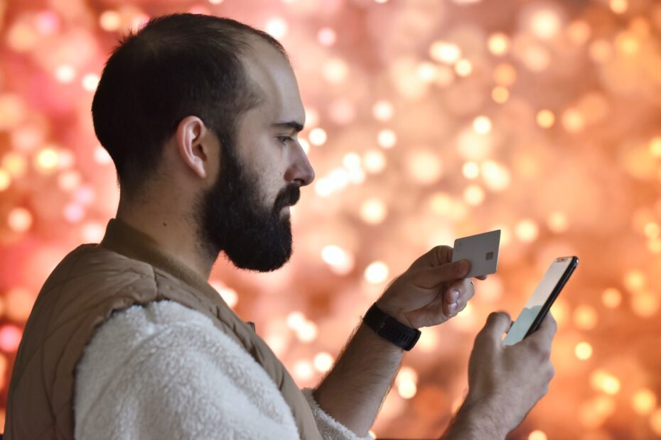 Homem barbudo segurando um cartão de crédito e um smartphone, realizando uma transação online, com fundo desfocado em tons quentes e brilhantes.