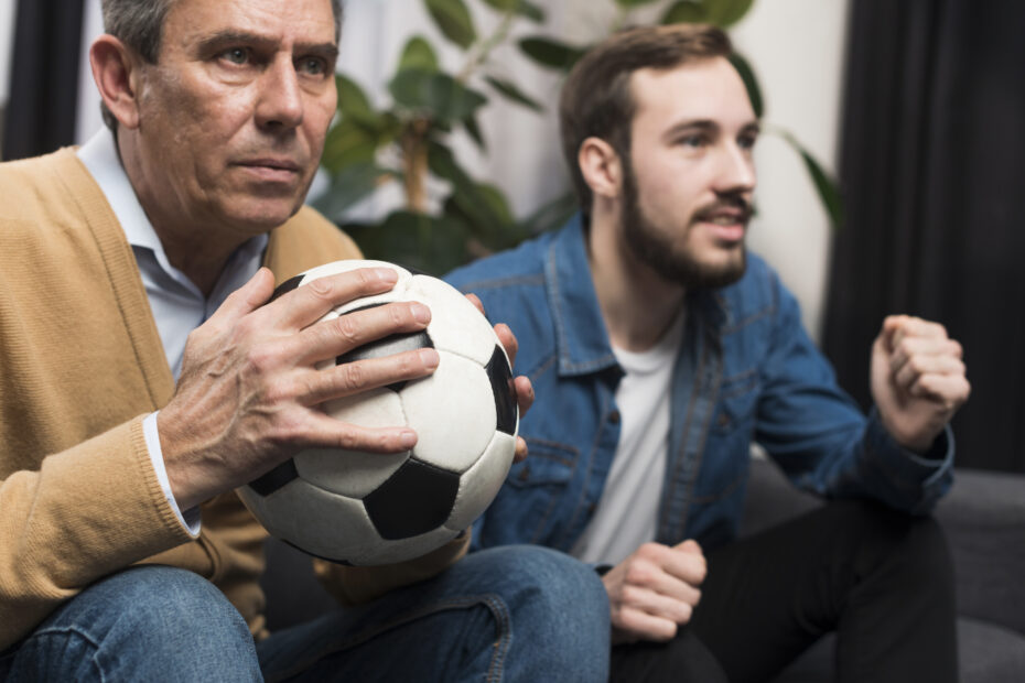 Dois homens assistem a uma partida de futebol com expressão de tensão e expectativa. O homem mais velho segura uma bola de futebol nas mãos, enquanto o mais jovem, ao lado, parece empolgado. Ambos estão sentados em um sofá em ambiente interno.