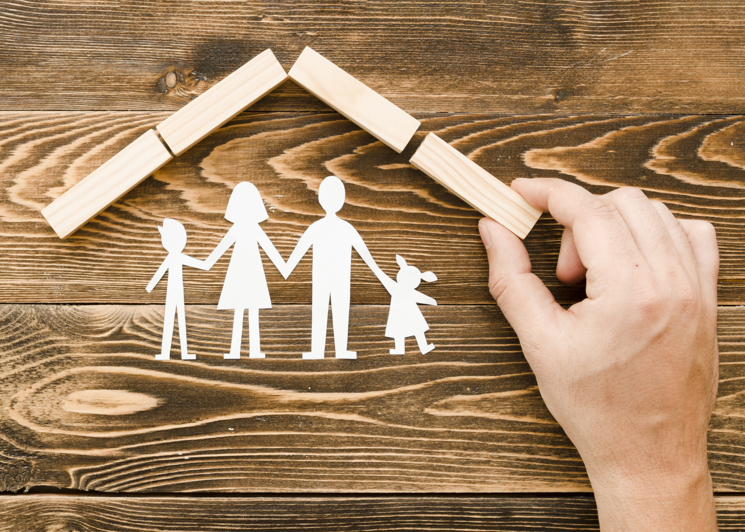 Representação de uma família feita com recortes de papel branco, composta por dois adultos e duas crianças, sob um telhado simbólico formado por blocos de madeira. Uma mão humana posiciona uma das peças do telhado sobre uma superfície de madeira.
