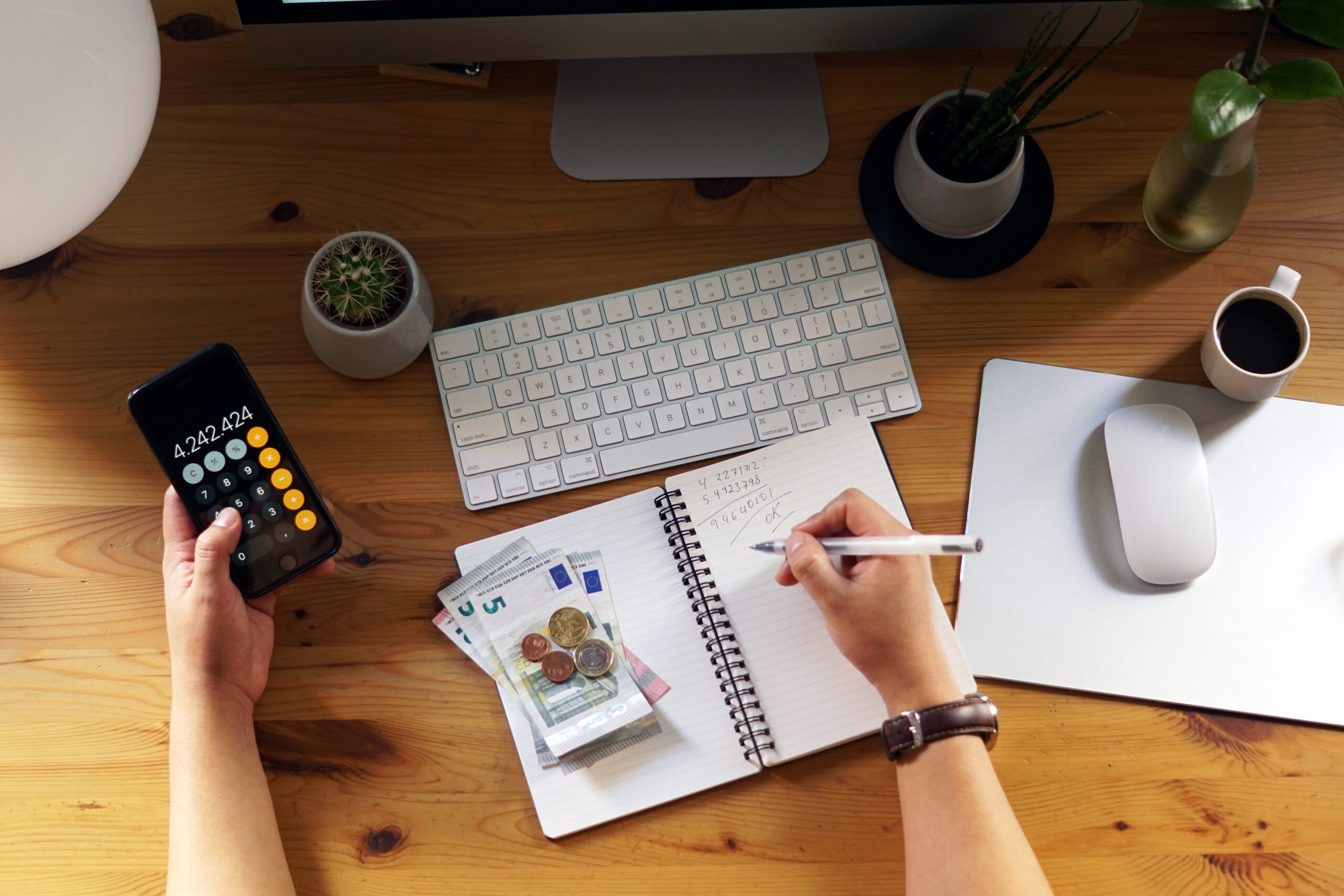 Pessoa sentada à mesa de trabalho usando uma calculadora no celular enquanto anota números em um caderno. Há cédulas e moedas de euro sobre o caderno, um teclado de computador, um mouse branco, um copo de café, plantas pequenas e objetos de escritório sobre a mesa de madeira.