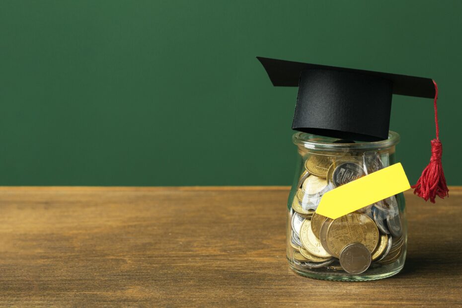 Pote de vidro cheio de moedas, com um chapéu de formatura preto e barbicacho vermelho apoiado em cima, simbolizando economia ou investimento em educação, sobre uma superfície de madeira com fundo verde.