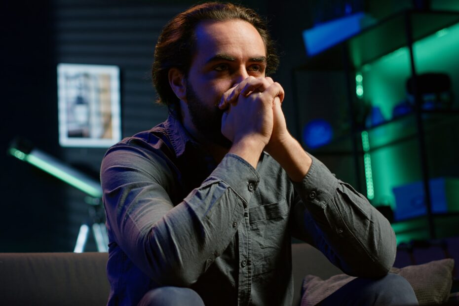 Homem jovem de barba e cabelo comprido preso, sentado em um ambiente escuro com iluminação azul e verde. Ele está com as mãos sobre a boca, em expressão de preocupação ou reflexão, olhando fixamente para frente. Ao fundo, há uma estante iluminada, objetos decorativos e um telescópio.