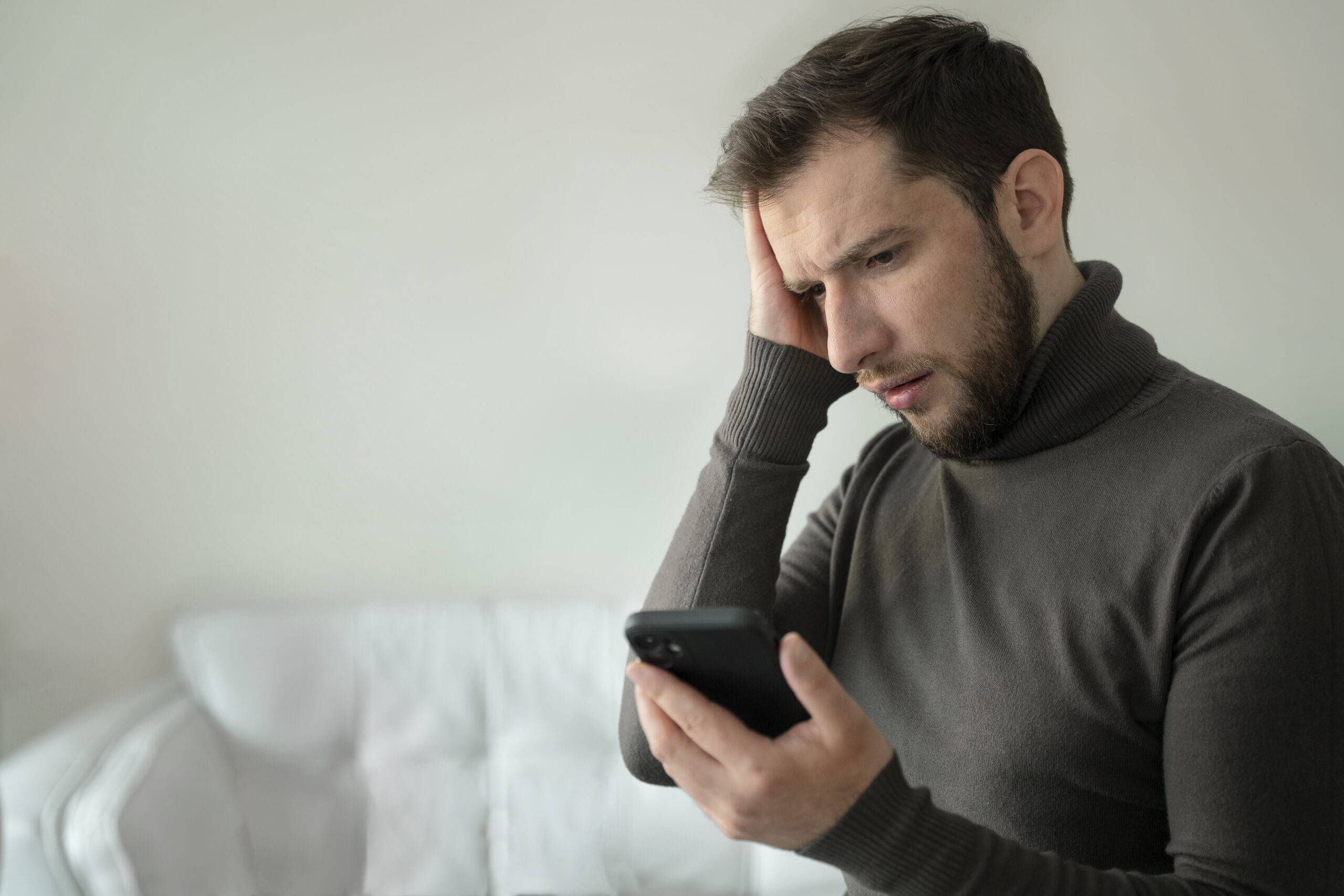 Homem jovem com expressão de preocupação olhando para o celular enquanto segura a cabeça, sentado em um ambiente interno com sofá branco ao fundo.